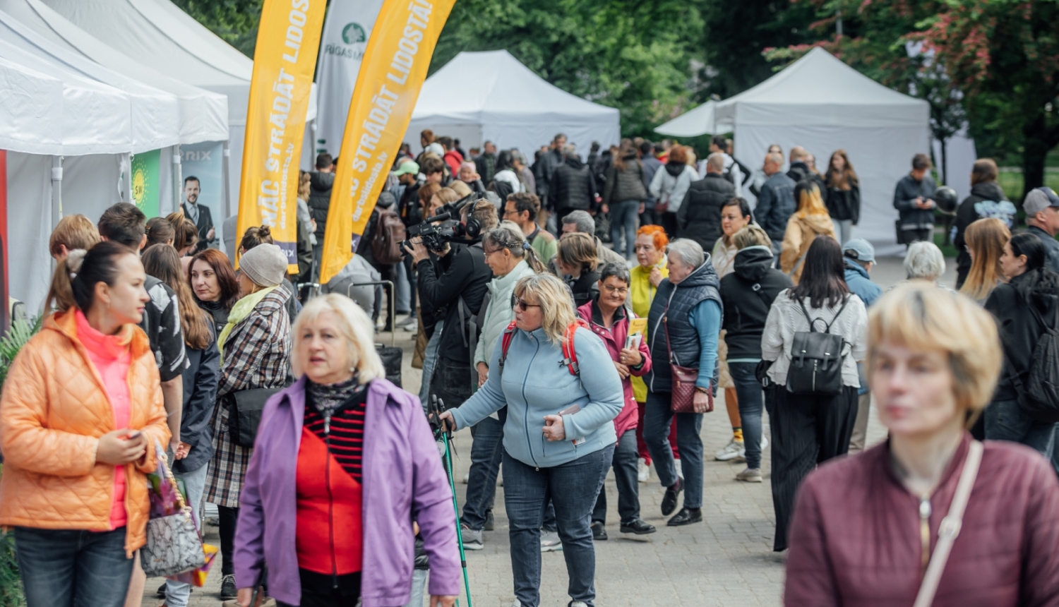 Pūlis ar cilvēkiem darba iespēju festivālā, malās baltas teltis un dzelteni vertikāli baneri