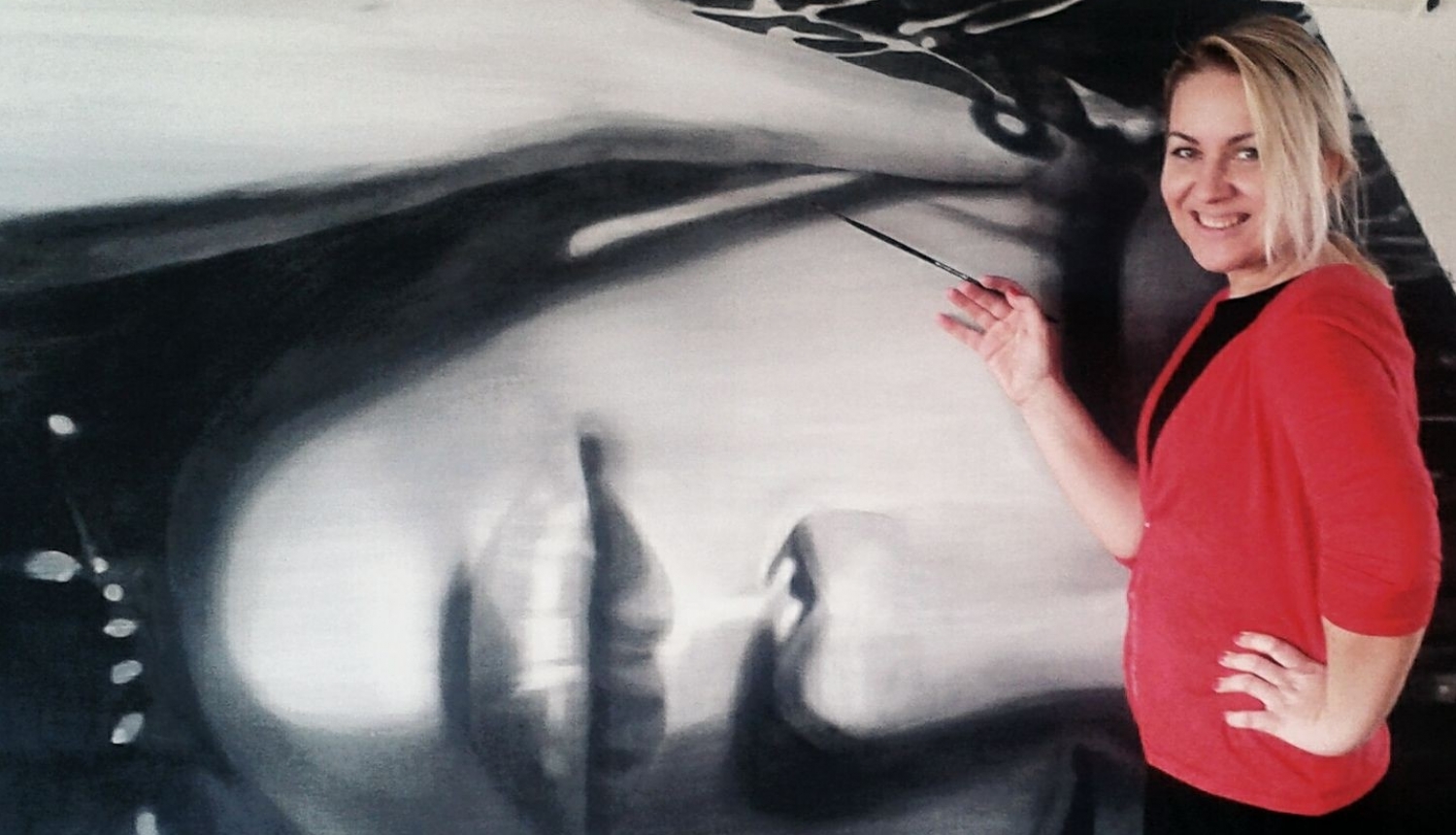 Māksliniece ar gaišiem matiem, sarkanā žaketē ar otu rokās pie lielizmērā mākslas darbu, kurā atspoguļots sievietes portrets